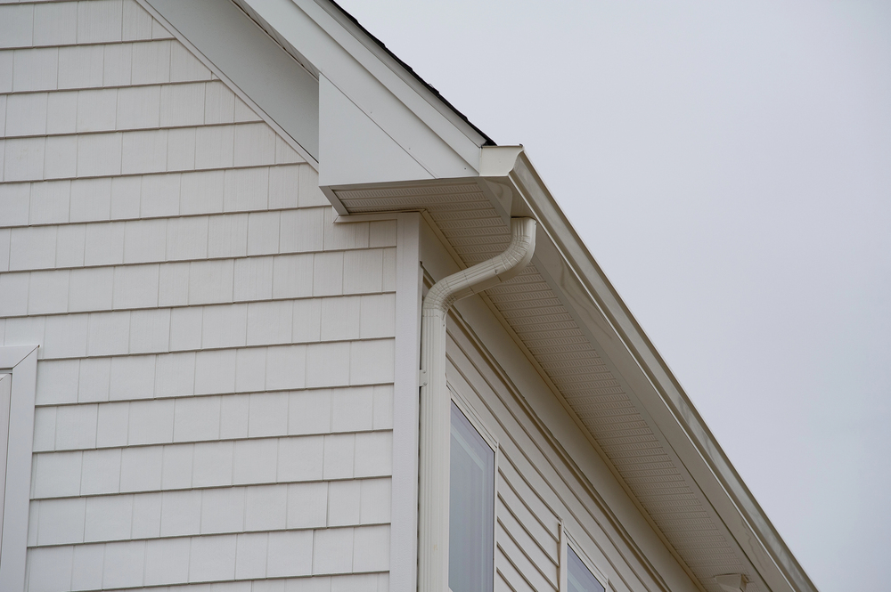 Close-up of a house exterior showing white siding, a rain gutter, and downspout against a cloudy sky.