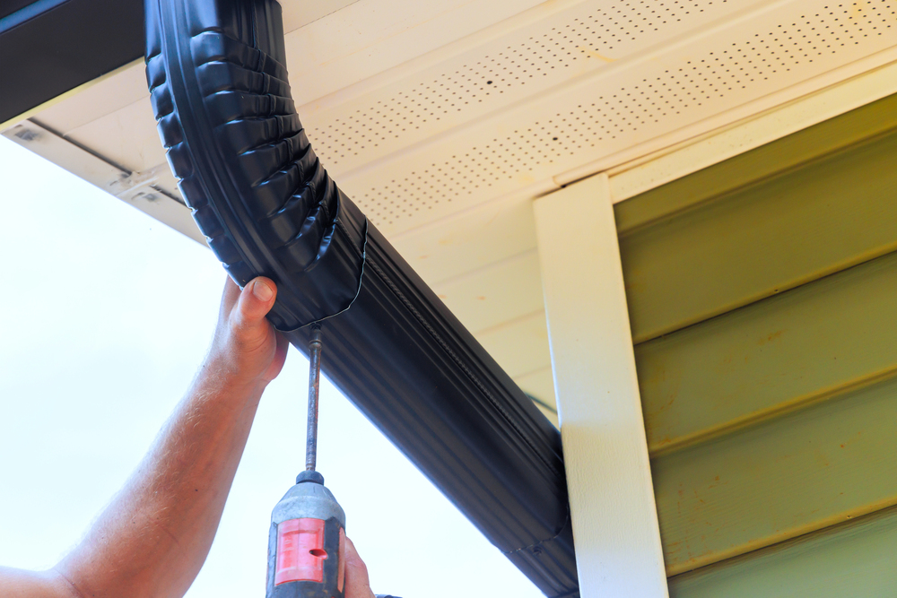 Person using a power drill to install or secure a black rain gutter onto the roof edge of a house with green siding.