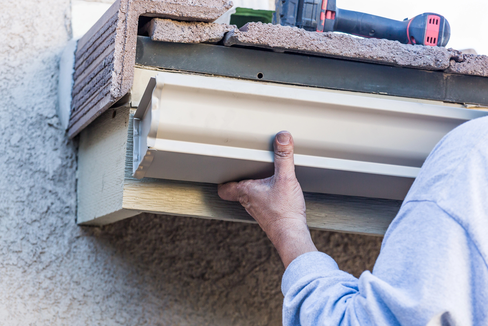 Person installing a white rain gutter under a roof edge, with a drill resting on top of the shingles.