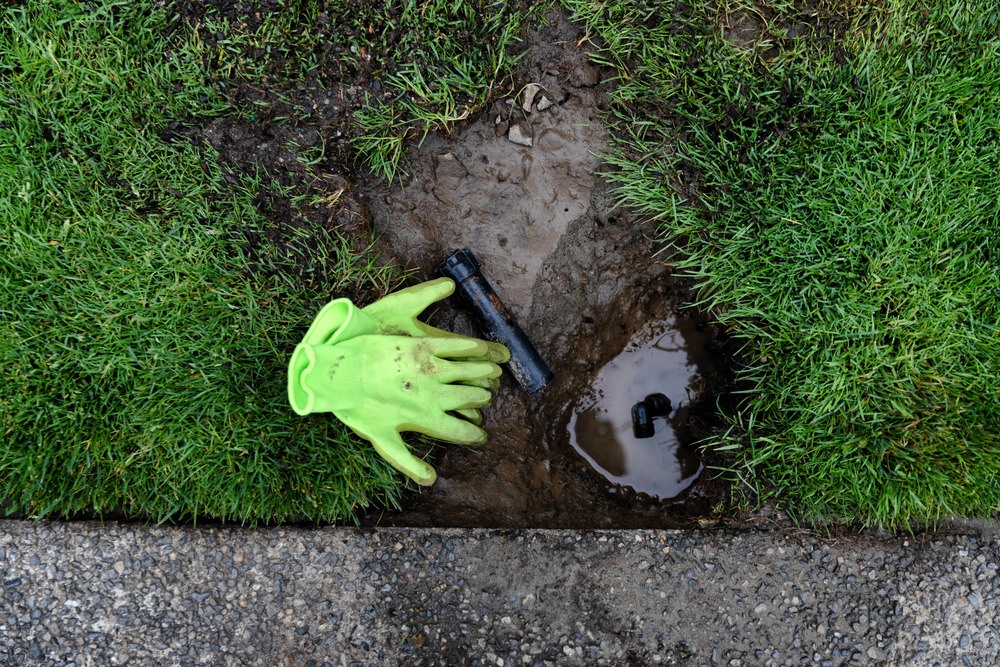 A muddy patch of grass shows a green work glove and exposed irrigation pipe near a concrete edge, suggesting ongoing sprinkler system repair.
