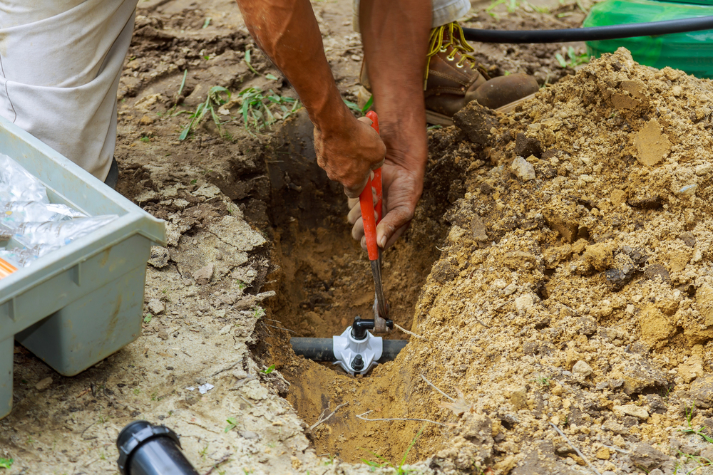 Person using a wrench to install or repair a sprinkler valve in a dirt trench, with a toolbox and tools nearby.